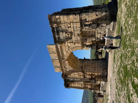       Two tourists stand near the grand stone arch of ancient Roman ruins under a bright blue sky
  