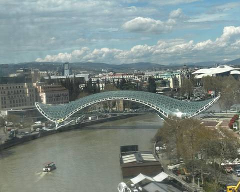       Modern arched glass pedestrian bridge spanning a river with Tbilisi cityscape and mountains in the background.
  