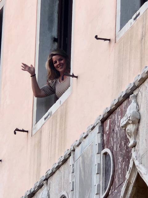       Smiling woman waving from an upper-storey window of a pastel stucco building
  