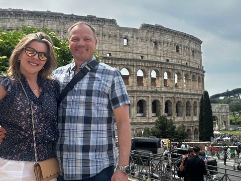       Smiling couple posing with the Colosseum towering behind them on a sunny afternoon in Rome
  