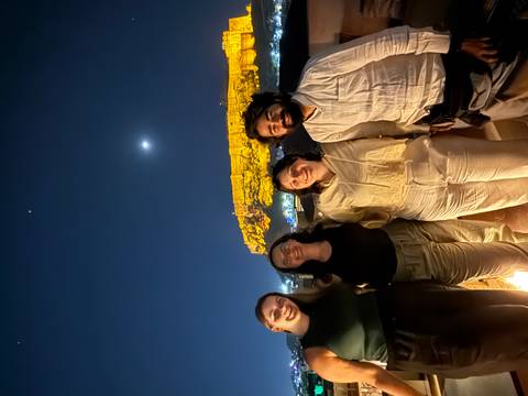       Four smiling friends pose at night with a brightly lit desert fortress and moonlit sky behind them.
  