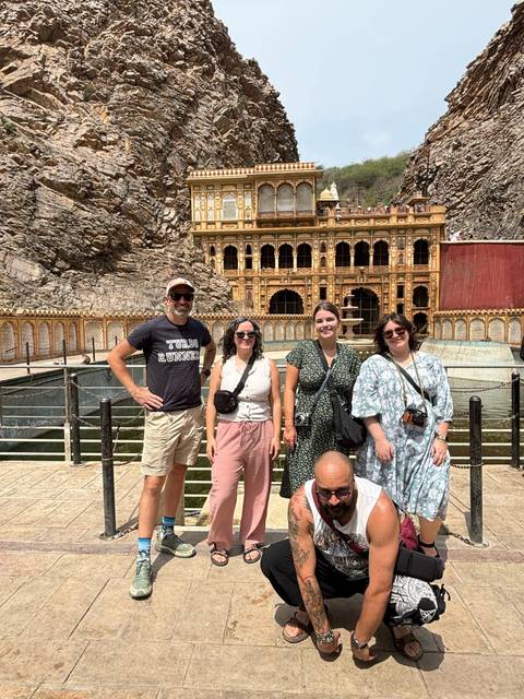       A travel group stands in front of a richly carved temple facade and sacred pool surrounded by rocky hills.
  
