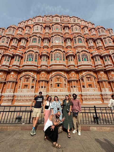       A group of travelers stands before the ornate red-sandstone façade of Jaipur’s Hawa Mahal under a clear sky.
  