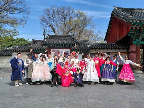       Large tour group dressed in vibrant traditional hanbok posing joyfully in front of an ornate palace gate.
  