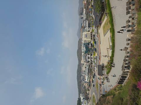       Wide portrait-orientation shot of a modern Korean town square with low buildings and distant hills beneath a hazy blue sky.
  