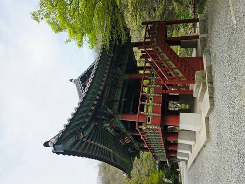       Colorfully painted wooden pavilion with ornate roof and red pillars set among greenery at a Korean temple site.
  