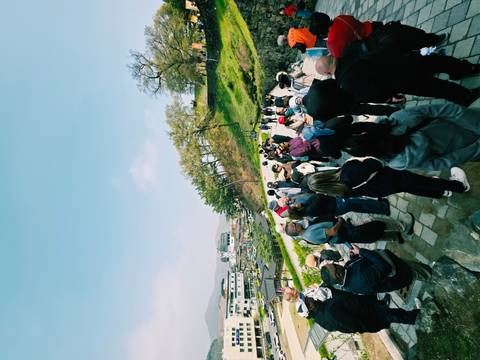       Large tour group walking along a cobbled path overlooking a hillside town and spring trees under blue skies.
  