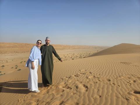       A couple poses atop rolling golden sand dunes under a clear sky in the desert.
  
