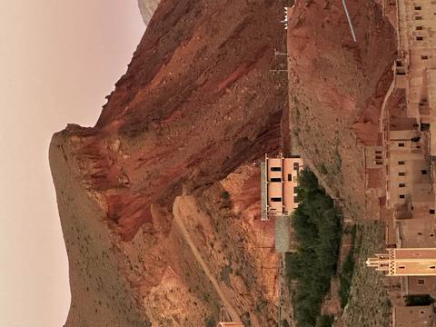       Reddish desert cliffs rise behind a small adobe village house at dusk light
  
