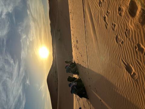       Camel riders rest on a high dune while the sun sets over rolling Sahara sand
  