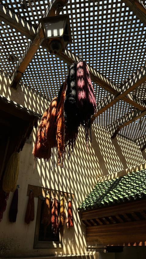       Bundles of dyed yarn hang from beams casting striped shadows below
  