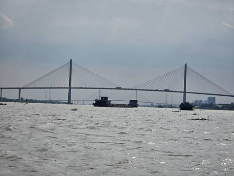       A cable-stayed bridge spans a wide, choppy river under a gray sky with barges passing below.
  