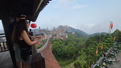       Visitors admire misty mountaintop castles and lush hills from a wooden balcony decorated with a red lantern.
  