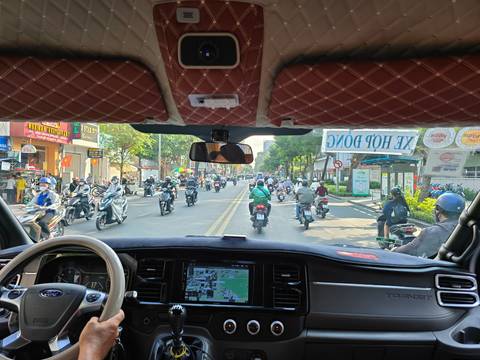       Street traffic of dozens of motorbikes and scooters viewed from inside a van’s windshield.
  
