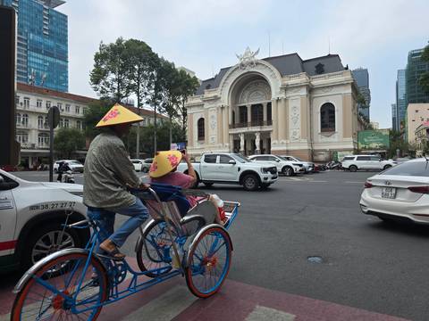       A traditional cyclo with passengers wearing conical hats passes the ornate Saigon Opera House.
  