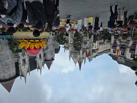       Crowds explore a European-style theme park square with stone castles and vibrant flower displays.
  