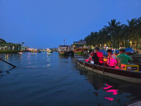       Colorful lantern-lit boats glide along a lively riverfront at blue hour with palm trees silhouetted.
  