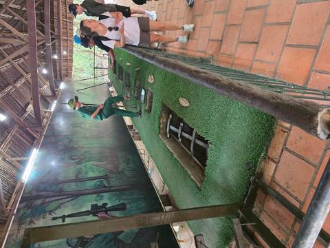       A guide demonstrates hidden spike traps at the Cu Chi Tunnels using a grass-covered display pit.
  