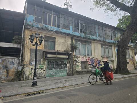       An older man pedals a bicycle past a weathered graffiti-covered colonial building.
  