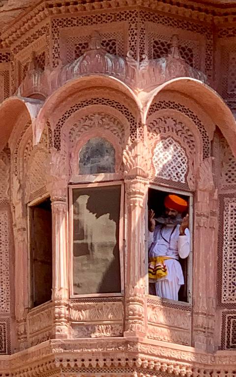       Man in orange turban peers from intricately carved pink sandstone window
  