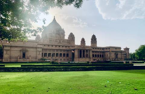       Grand sandstone palace with domes and turrets set behind manicured green lawns under a bright sky.
  