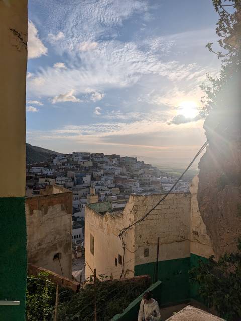       View through ruined walls toward a hillside town bathed in late-day sun with distant plains beyond.
  