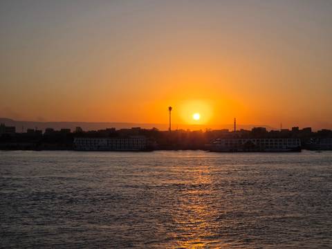       Orange sun setting behind silhouette of riverside cityscape along wide calm river.
  
