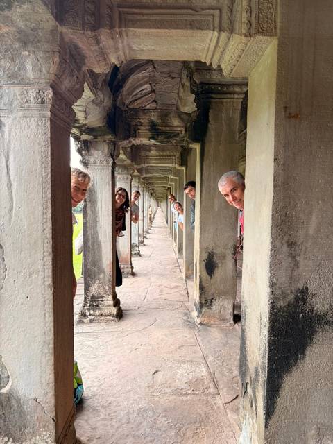       Smiling travellers peek out between stone columns in a long Angkor temple corridor.
  