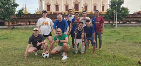       Travellers and local youths pose with football on grassy field beside ornate stupa.
  