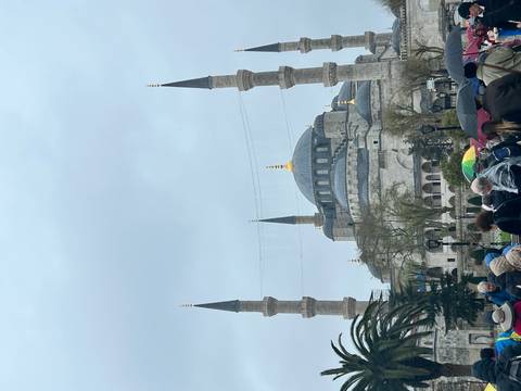       View of the Blue Mosque with crowds and umbrellas under an overcast sky.
  
