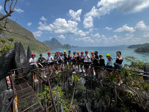       Helmeted travellers stand on a jagged limestone viewpoint overlooking turquoise sea and dramatic karst islands.
  
