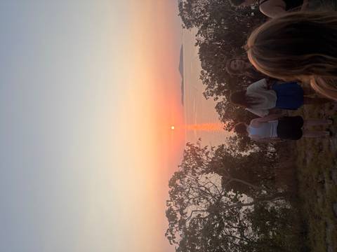       Travellers watch an orange sun set over calm sea from a grassy hilltop.
  