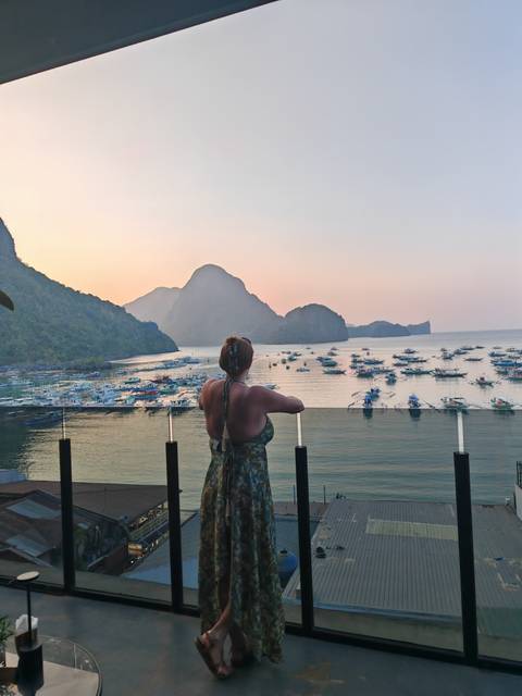       Traveller enjoys pastel sunset views of El Nido’s bay and karst formations from a glass balcony.
  
