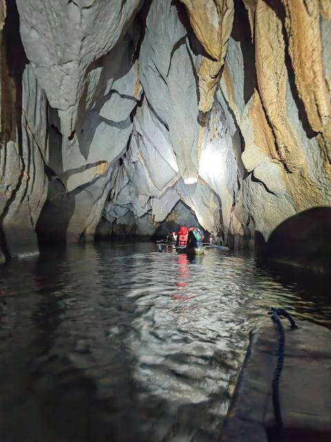       Small guided boat drifts through dramatic rock chambers of the subterranean river cave, helmets lit by torches.
  