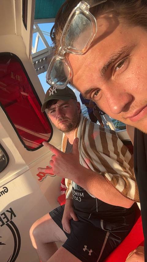       Close-up boat selfie of two young men wrapped in a towel with sea and sunset light in background.
  
