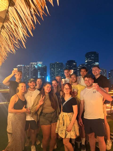       Group of travellers raise cups on a rooftop at night with Manila’s illuminated skyline behind.
  