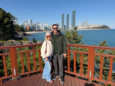       Two travellers smile on a wooden lookout with Busan’s skyscraper-lined coastline behind.
  