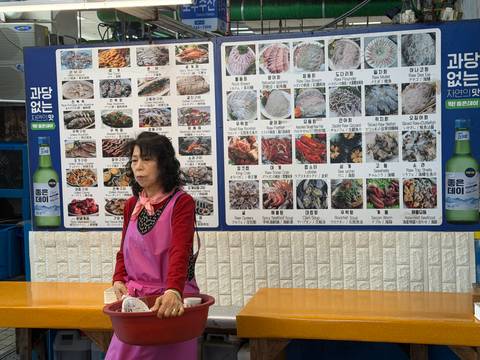       Market vendor in apron waits before colourful seafood photo menus on a wall.
  