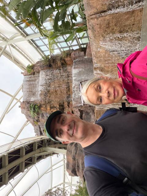       Selfie of two travellers in front of an indoor tiered rock waterfall feature.
  