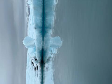       Solitary iceberg reflects in still milky-blue glacial water under grey skies.
  