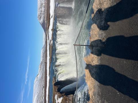       Mighty Godafoss waterfall thunders over a horseshoe cliff as onlookers’ shadows fall in foreground.
  