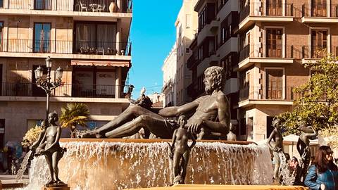       Classical fountain with reclining statue surrounded by cherubs in a lively city square
  