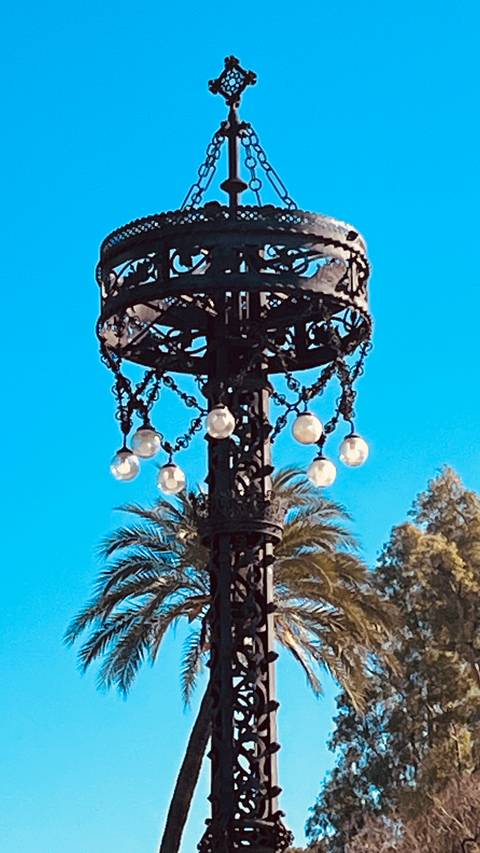       Close-up of a decorative wrought-iron streetlamp with hanging glass bulbs against a bright blue sky and palm trees.
  