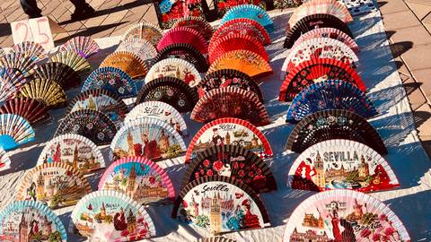       Display of colorful hand fans with Sevilla motifs arranged on a market blanket.
  