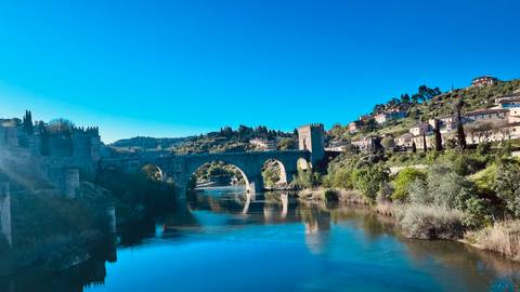       Stone medieval bridge spanning a calm river with hillside town and greenery in the background.
  