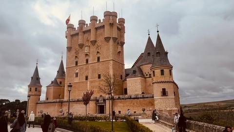       Large fairytale stone castle with conical towers under cloudy skies, visitors walking around the courtyard.
  