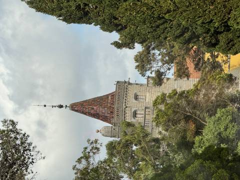      A pointed tiled turret rises above lush trees under cloudy skies, Sintra, Portugal.
  