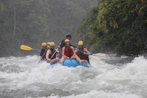      Raft with excited paddlers plunging through white-water rapids amid jungle river scenery.
  