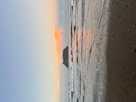       Island silhouette under pastel sunset skies reflected on calm coastal waters from a sandy shoreline.
  