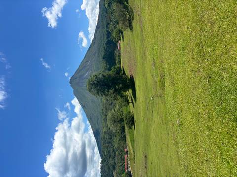       Bright midday view of conical Arenal Volcano framed by grassy meadow and scattered clouds.
  